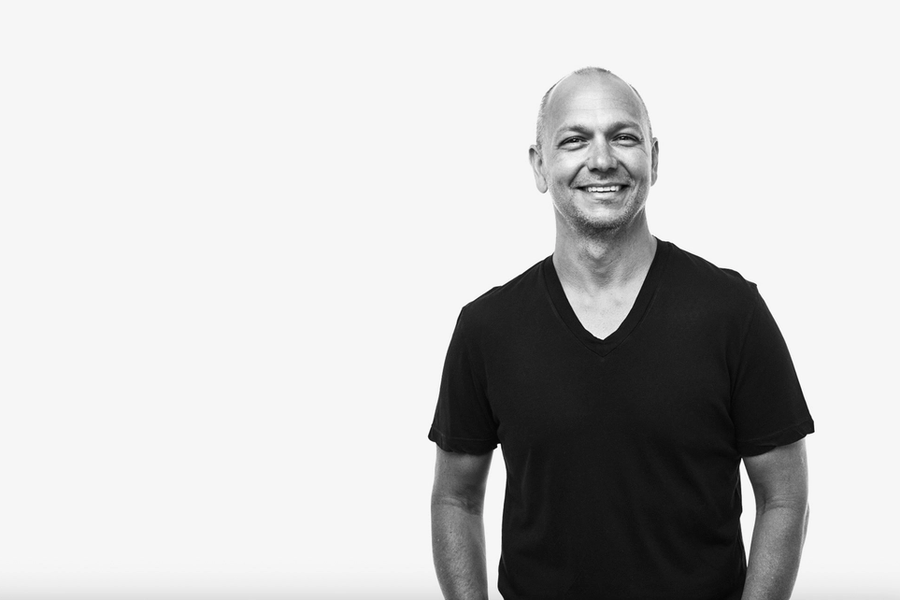 A black-and-white photo of Tony Fadell, smiling, wearing a short-sleeved, V-neck T-shirt. He is standing casually with one hand in his pocket against a plain white background.